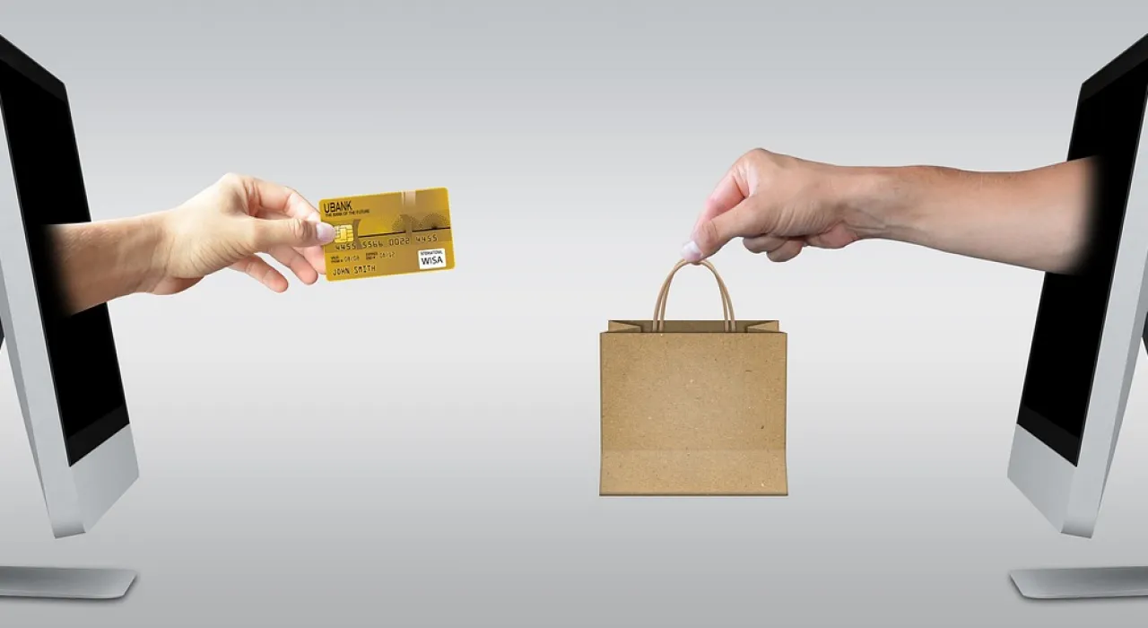 imagen de dos ordenadores desde los que sale de uno una mano con una tarjeta de credito y del otro otra mano con una bolsa